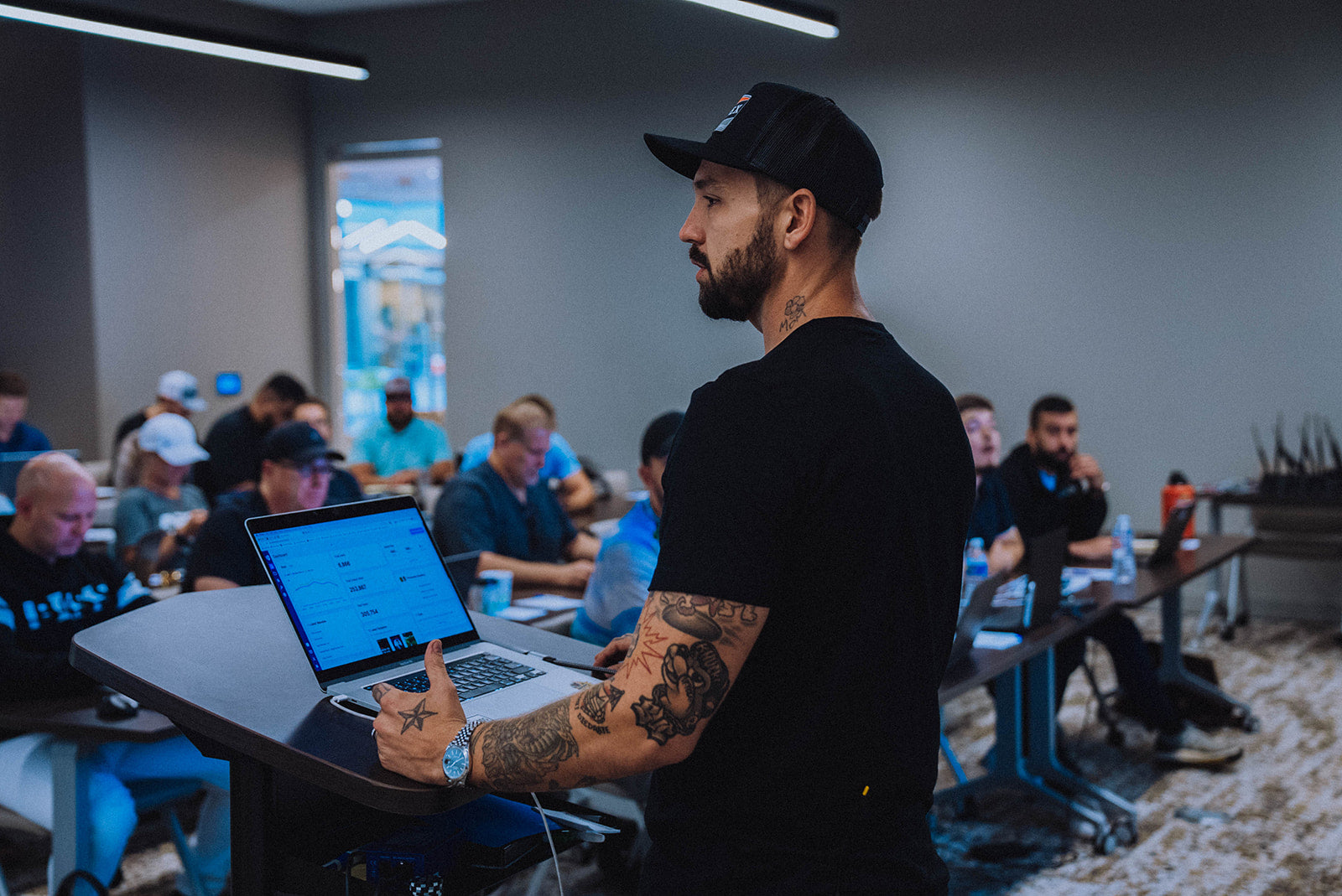 A man with tattoos and a beard, wearing a black t-shirt and a black baseball cap, stands at a lectern using a laptop in a presentation or lecture setting, with an audience seated in the background.
